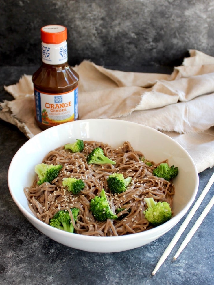 Bowl of soba noodles with broccoli and creamy dressing with chopsticks in foreground, bottle of Soy Vay marinade in background with cloth.