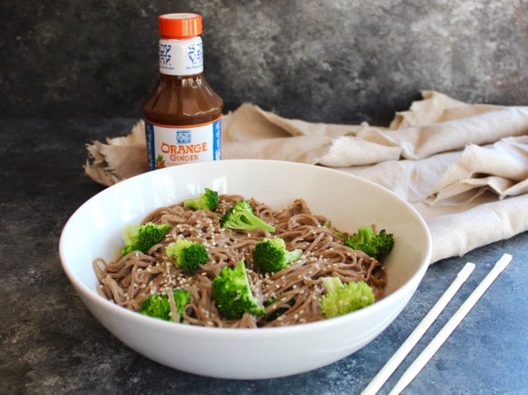 Bowl of soba noodles with broccoli and creamy dressing with chopsticks in foreground, bottle of Soy Day marinade in background with cloth.