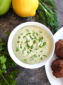 White bowl of herb tahini sauce topped with chopped fresh parsley alongside a falafel plate, fresh parsley, fresh dill, lemon and lime on the side.