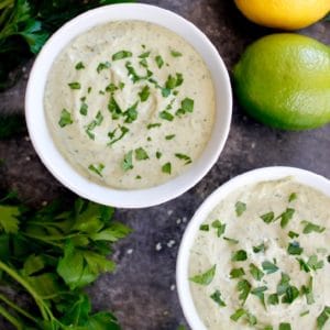 Two white bowls of herb tahini sauce topped with chopped parsley on concrete background with fresh parsley, lemon, lime and sesame seeds.