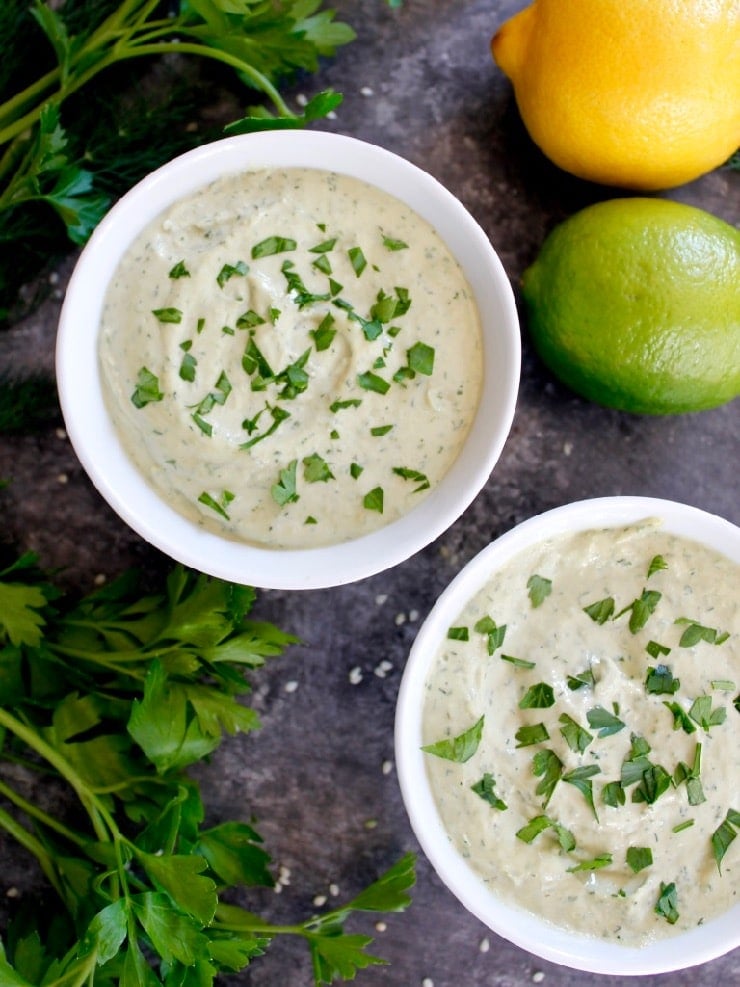 Two white bowls of herb tahini sauce topped with chopped parsley on concrete background with fresh parsley, lemon, lime and sesame seeds.