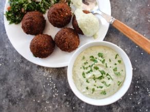 White bowl of herb tahini sauce topped with chopped fresh parsley alongside a falafel plate. One piece of falafel topped with tahini sauce, spoon beside it. Tabouli salad in background.