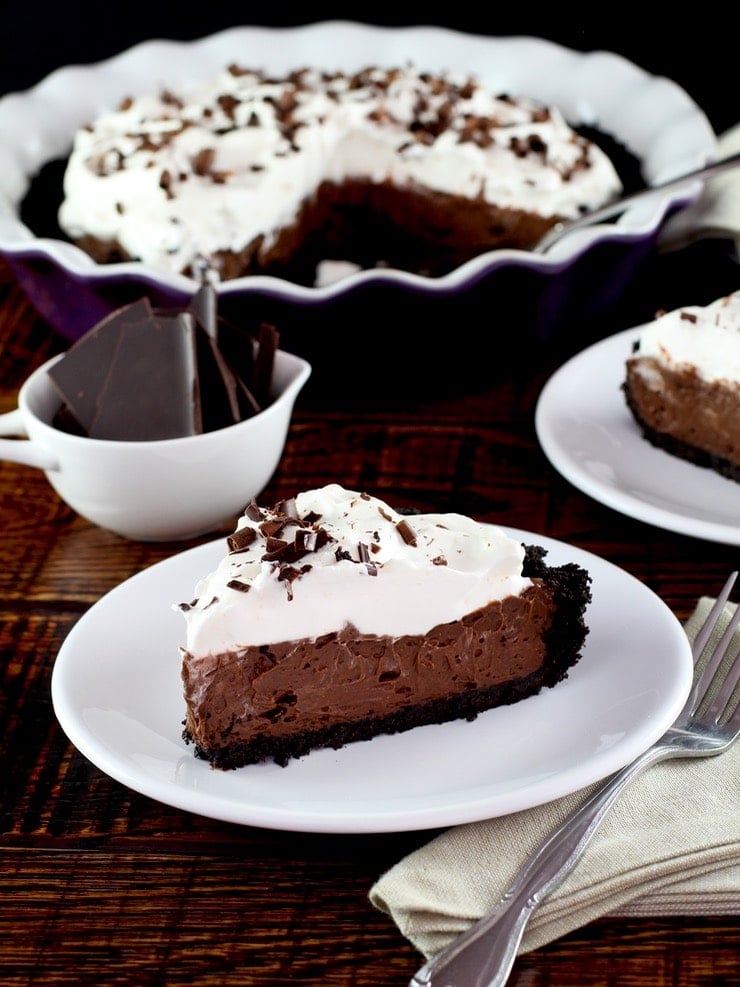 Slice of chocolate pie topped with whipped cream and chocolate shavings on a white plate. There is a whole pie in the background.