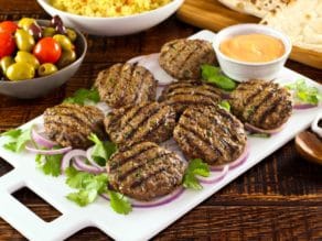 White platter of Middle Eastern Burgers char-grilled on a bed of purple onion rings and herbs, with spicy sriracha mayo sauce on the side, dish of yellow couscous, pita bread and olives in background.