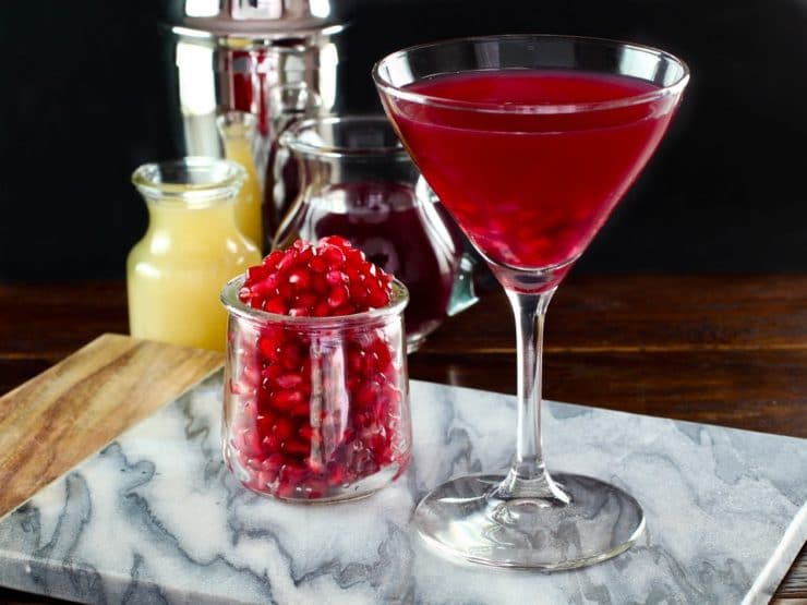 A bright red cocktail in a martin-style cocktail glass next to a small jar of pomegranate seeds