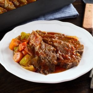 A slice of beef brisket next to a side of vegetables and topped with gravy on a round white plate.