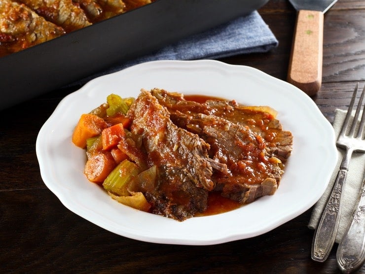 A slice of beef brisket next to a side of vegetables and topped with gravy on a round white plate.