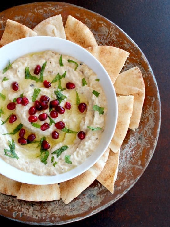 Vertical shot of Roasted Eggplant Yogurt Dip topped with pomegranate arils and fresh mint, on a platter with pieces of pita.