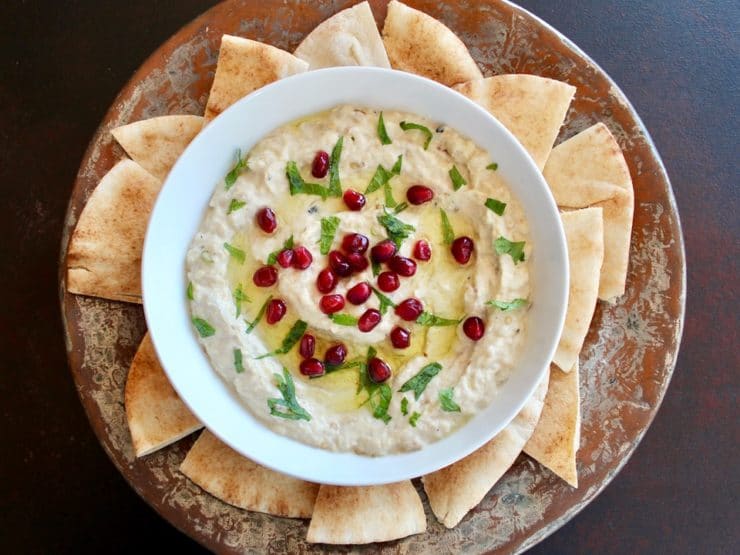 Horitzontal shot of Roasted Eggplant Yogurt Dip topped with pomegranate arils and fresh mint, on a platter with pieces of pita.
