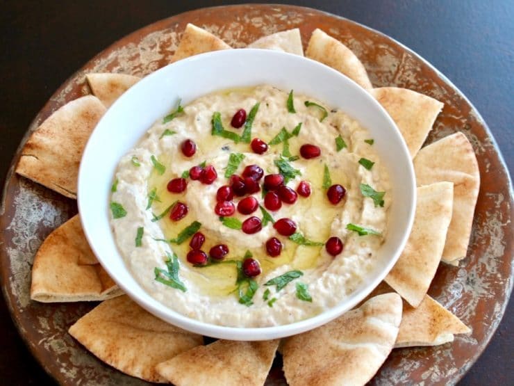 Overhead shot of Roasted Eggplant Yogurt Dip topped with pomegranate arils and fresh mint, on a platter with pieces of pita.