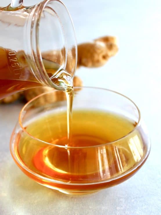 Vertical shot of glass carafe pouring agave ginger syrup into bowl, whole ginger root in background.