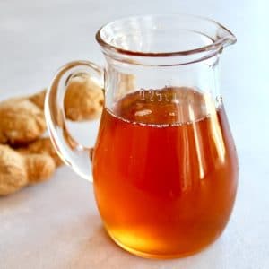 Glass carafe of ginger agave syrup on concrete background with whole ginger root behind.