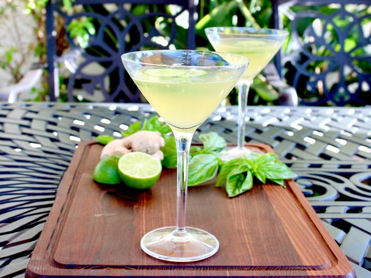 Two cucumber ginger martinis in martini glasses on wooden cutting board garnished with cucumber slices, basil, lime and whole ginger root in background, on outdoor black table. Horizontal shot.