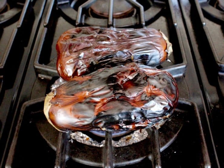 Two flame roasted eggplants with charred skin on stovetop grate, gas stove.