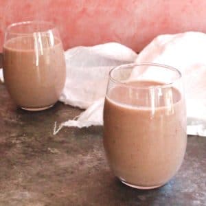 Two glasses of acai almond milk smoothie on grey concrete table with white cloth, pink wall in background.