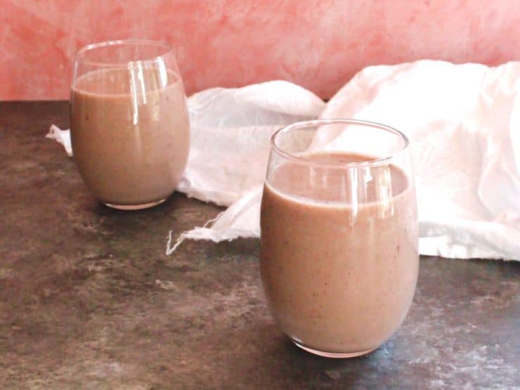 Two glasses of acai almond milk smoothie on grey concrete table with white cloth, pink wall in background.