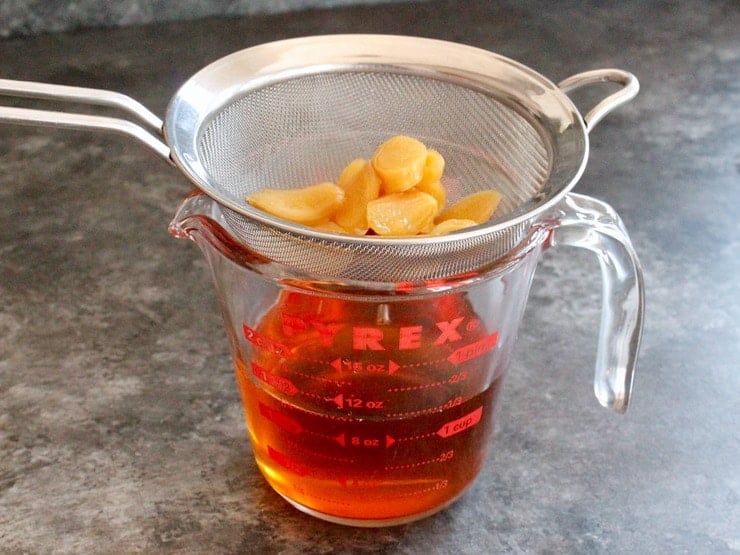 Straining agave syrup into glass measuring cup, sliced ginger in mesh strainer above.