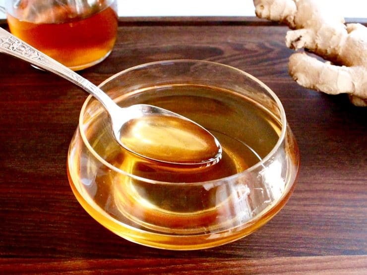 Agave ginger syrup in bowl, antique teaspoon spooning out syrup, on wooden cutting board with ginger and syrup carafe in background.