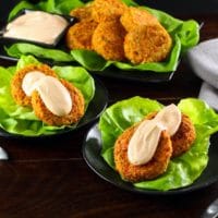 Two small dishes holding two salmon cakes topped with a spicy Sriracha sauce on a bed of lettuce. A platter of fish cakes and sauce is in the background.