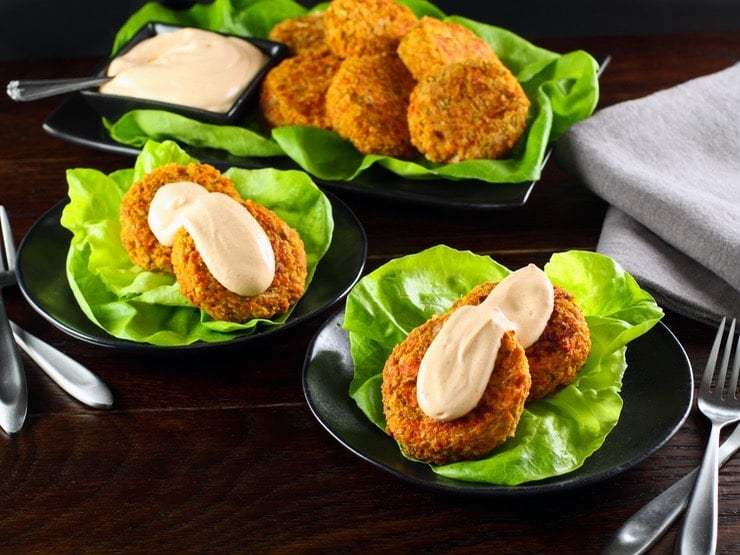 Two small dishes holding two salmon cakes topped with a spicy Sriracha sauce on a bed of lettuce. A platter of fish cakes and sauce is in the background.