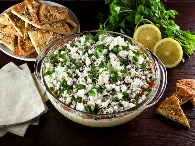 A large glass serving bowl filled with a layered dip that includes hummus, greek yogurt, Israeli salad, olives, feta cheese, and cilantro. A dish of pita chips is sitting off to the side.