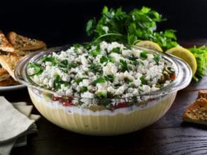 A large glass serving bowl filled with a layered dip that includes hummus, greek yogurt, Israeli salad, olives, feta cheese, and cilantro.