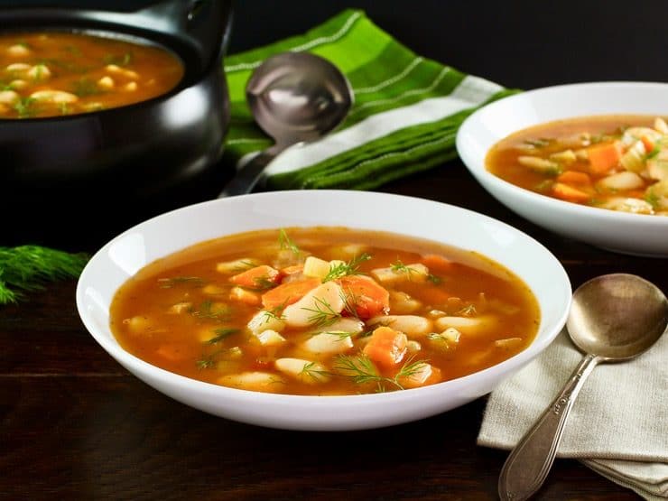 A bowl of butter bean soup in a red broth in a white soup bowl. A black pot filled with soup sits in the background next to a ladle on top of a white and green striped towel.