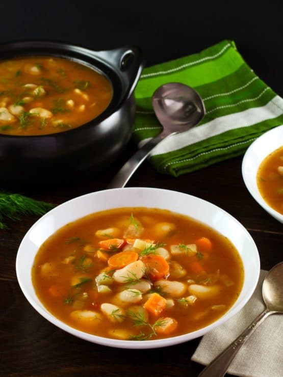 A bowl of butter bean soup in a red broth in a white soup bowl. A black pot filled with soup sits in the background next to a ladle on top of a white and green striped towel.