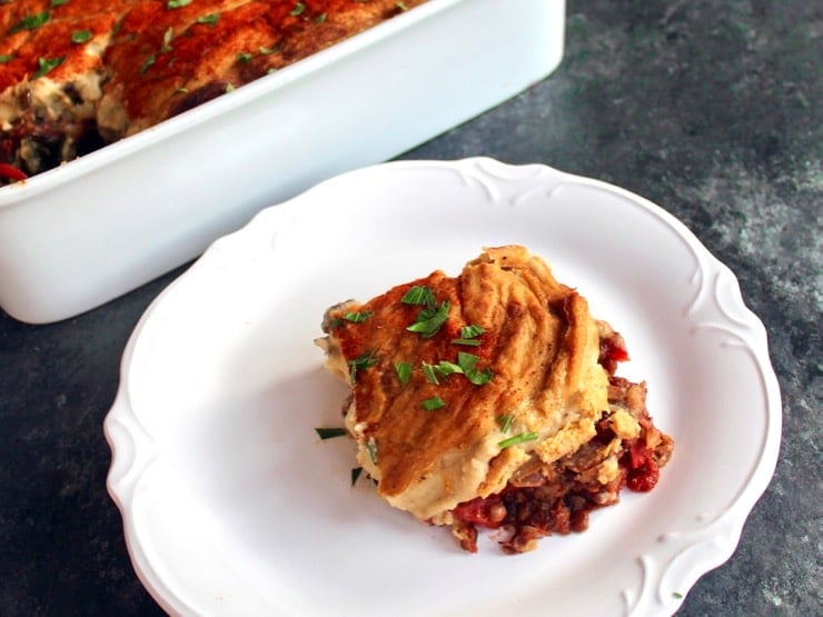 Slice of Hummus Baked Vegan Moussaka on a white plate with scalloped edges, casserole dish in background, on a grey concrete surface.