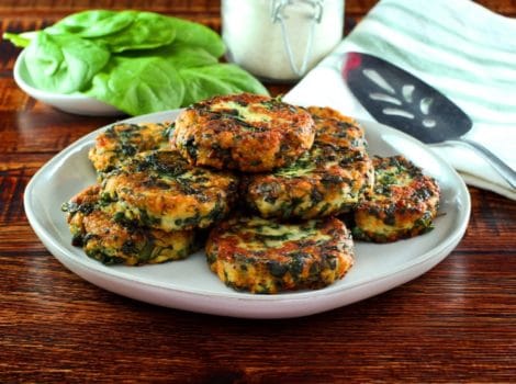 Horizontal shot - plate of fried keftes de espinaca - spinach keftes - piled on a white plate with fresh spinach, canister of breadcrumbs, spatula and cloth napkin in background.