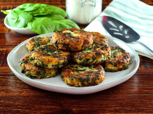 Horizontal shot - plate of fried keftes de espinaca - spinach keftes - piled on a white plate with fresh spinach, canister of breadcrumbs, spatula and cloth napkin in background.