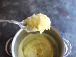 Spoon scooping up creamy mashed potatoes from a pot on concrete countertop.