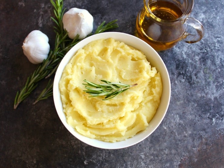 Overhead shot - bowl of creamy golden mashed potatoes topped with sprig of fresh rosemary, carafe of olive oil on the right, heads of garlic with rosemary sprig on the left.
