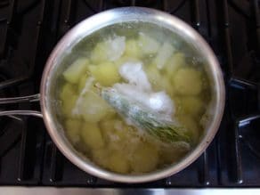 Pot of simmering potato chunks with cheesecloth bundle of rosemary floating on top on stovetop.