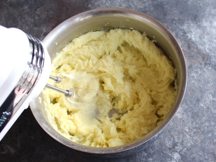 Hand mixer whipping mashed potatoes to a smooth creamy texture in a stainless steel mixing bowl.