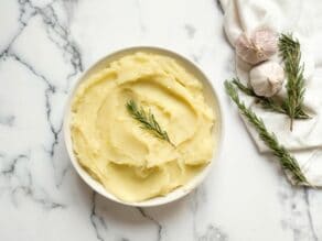 Overhead shot - Dish of olive oil mashed potatoes garnished with fresh rosemary on a marble countertop, garlic heads and rosemary in background with linen towel.