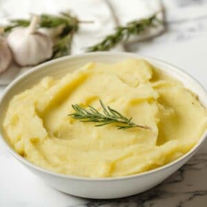 Horizontal shot - Dish of olive oil mashed potatoes garnished with fresh rosemary on a marble countertop, garlic heads and rosemary in background with linen towel.