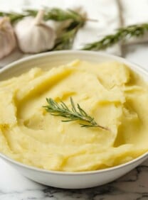 Square featured shot - Dish of olive oil mashed potatoes garnished with fresh rosemary on a marble countertop, garlic heads and rosemary in background with linen towel.