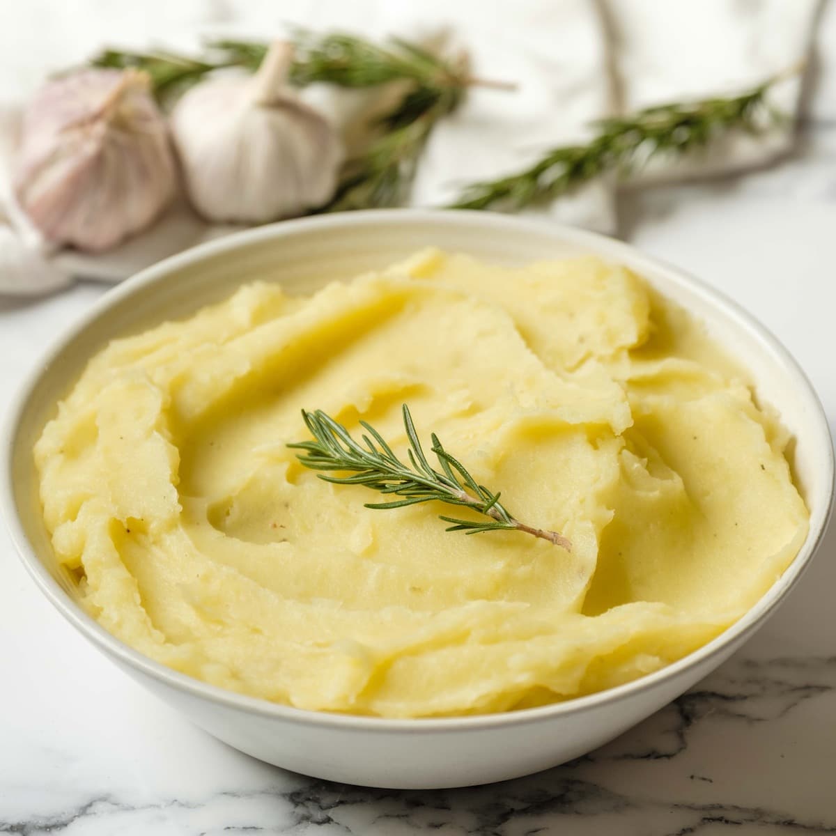Square featured shot - Dish of olive oil mashed potatoes garnished with fresh rosemary on a marble countertop, garlic heads and rosemary in background with linen towel.