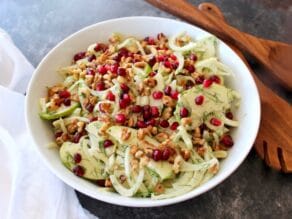 A bowl of Fennel Apple Salad with Cider Tahini Dressing, topped with pomegranate seeds, with wooden utensils and a white napkin.