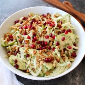 A bowl of Fennel Apple Salad with Cider Tahini Dressing, topped with pomegranate seeds, with wooden utensils and a white napkin.