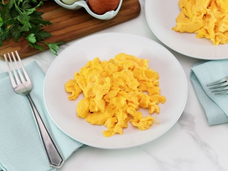 Fluffy scrambled eggs on a plate with fork and cloth napkin, on a marble countertop. Wooden cutting board with egg carton - brown and white eggs, and fresh parsley. Another plate of scrambled eggs in background.