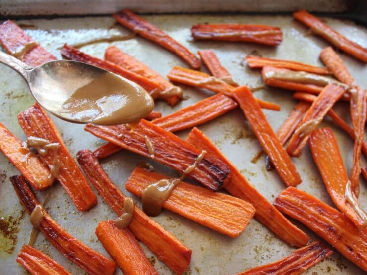 Spoon drizzling roasted carrots with tahini glaze on baking sheet.