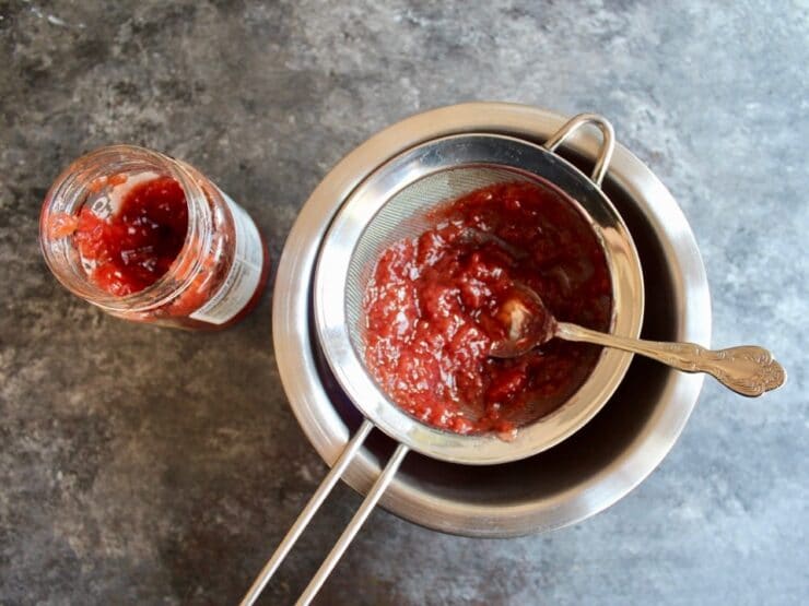 Jam being strained in a wire mesh strainer with spoon over a bowl on a concrete background, jar of jelly on the side.