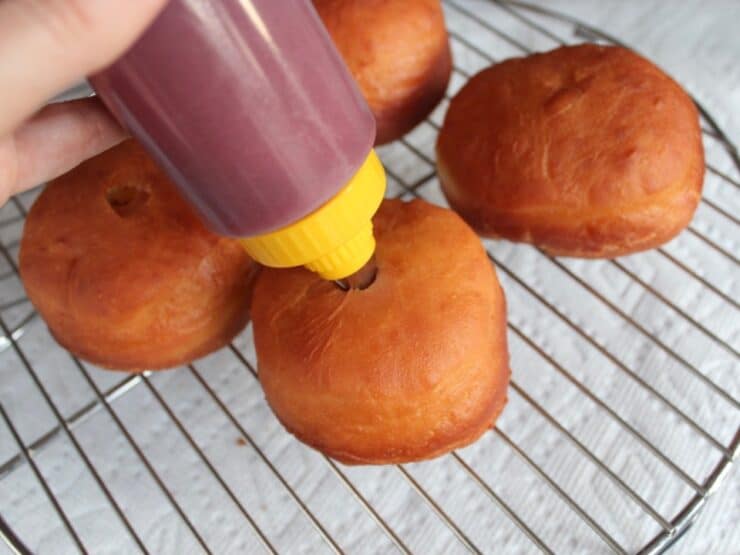 Sufganiyot on a wire rack. A bottle of filling with a pastry tip is inserted into one sufganiyot.
