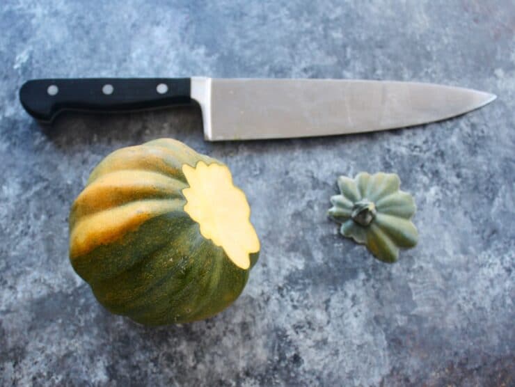 Acorn squash, stem end sliced off, stem end beside it with chef's knife above on concrete surface