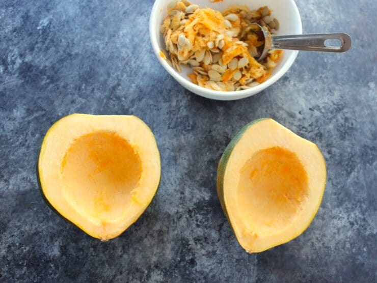 Two acorn squash halves with bowl of scooped seeds and pulp, tablespoon in background