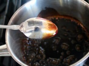 Spoon upside down showing thickened balsamic coating the back of the spoon, small saucepan with bubbling balsamic reduction sauce on stovetop in background