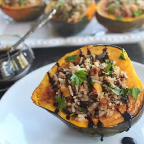 Horizontal shot of Vegan Stuffed Acorn Squash drizzled with balsamic reduction on small plate in foreground with glass of balsamic reduction and tray of stuffed squash in background