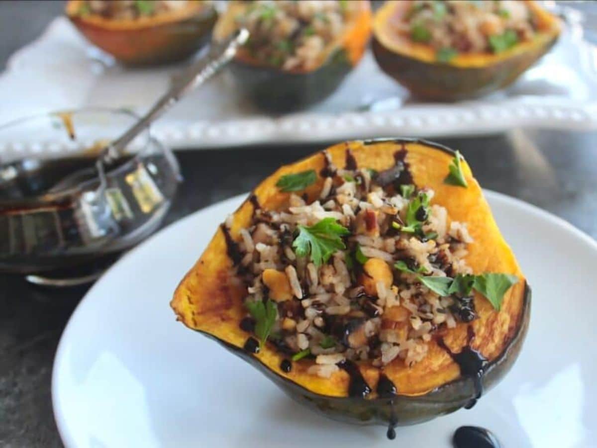 Horizontal shot of Vegan Stuffed Acorn Squash drizzled with balsamic reduction on small plate in foreground with glass of balsamic reduction and tray of stuffed squash in background